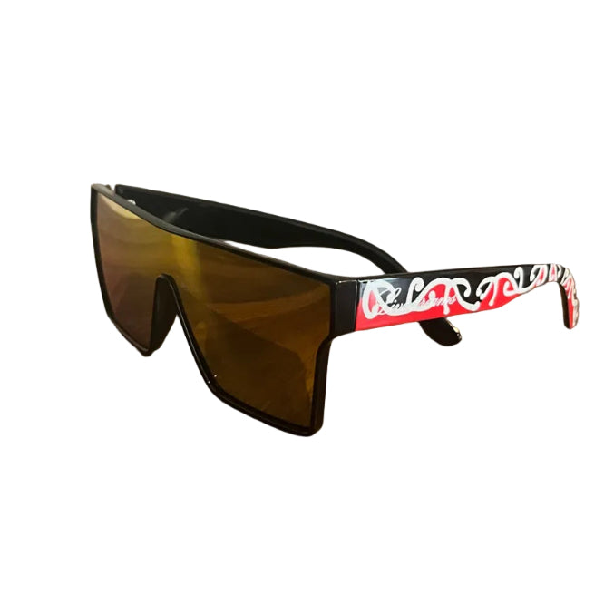 A pair of wraparound shield-style sunglasses with black frames and amber-tinted lenses, resting on a wooden table. The left temple arm features a red, white, and black graphic pattern with stylized lettering.