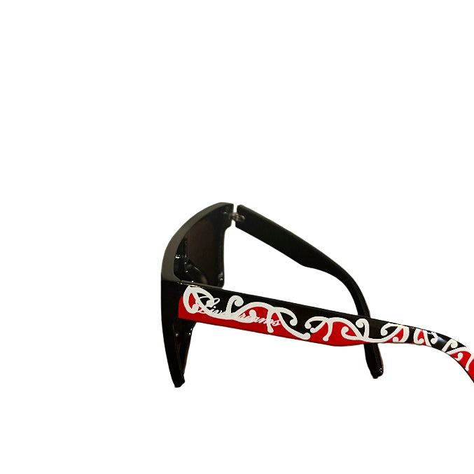 Black sunglasses with a red and white patterned temple arm, resting on a wooden table against a beige wall.