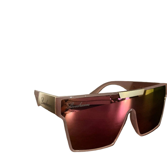 A pair of oversized shield sunglasses with a translucent pink frame and mirrored pink lenses, resting on a dark wooden table with a blurred indoor background.