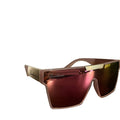 A pair of oversized shield sunglasses with a translucent pink frame and mirrored pink lenses, resting on a dark wooden table with a blurred indoor background.